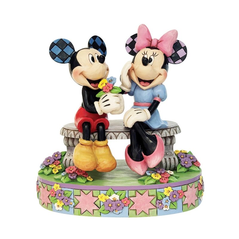Jim Shore-beeldje van Mickey die Minnie een boeket bloemen geeft op een parkbankje, omringd door kleurrijke bloemen en versierd met rosemaling- en quiltpatronen.